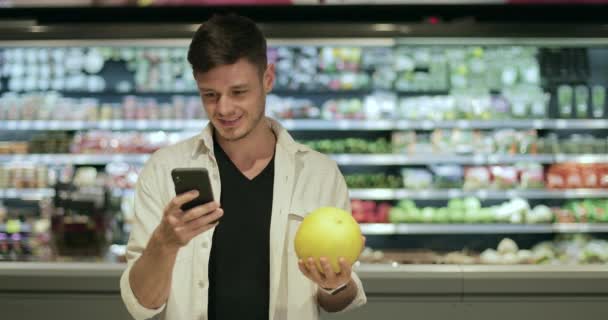 Lachende man die pomelo vasthoudt en smartphone gebruikt terwijl hij naar het scherm kijkt in de supermarkt. Millennial knappe man staande en het houden van fruit tijdens het winkelen. Wazige achtergrond. — Stockvideo