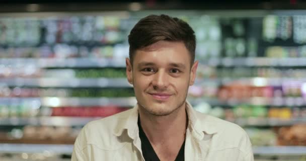 Портрет веселого хлопця, який дивиться на камеру, стоячи в супермаркеті. Близький погляд на щасливого тисячолітнього чоловіка, який позує і посміхається. Концепція людей і спосіб життя. Розмитий фон . — стокове відео