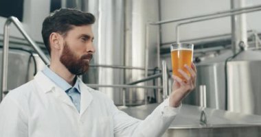 Dikkat çeken erkek işçinin cam bardakta taze yapılmış içeceğe bakışı. Modern bira fabrikasında dikilirken beyaz ceketli yakışıklı adam bira kalitesini ölçüyor..
