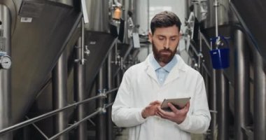 Beyaz laboratuvar önlüklü sakallı adam bira fabrikasındaki tablete veri giriyor. Yürürken ve otomatik içecek ekipmanlarına bakarken dijital ekrana dokunup daktilo eden dikkatli bir erkek işçi..