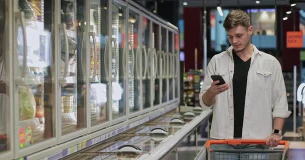 Giovane bel ragazzo guardando lo schermo dello smartphone e cercando prodotto nel reparto congelato del negozio. Millennial uomo utilizzando il telefono e camminando con carrello vicino supermercato frigoriferi . — Video Stock