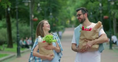 Parkta alışveriş yaptıktan sonra güler yüzlü bir çift konuşuyor ve gülüyor. Güzel bir kadın ve erkek sağlıklı yiyecek ve gülümseyen kağıt torbalar taşıyorlar..