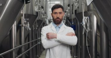 Kameraya bakıp el ele tutuşan erkek işlemci görüntüsü. Beyaz laboratuvar önlüklü sakallı adam çelik fıçıların önünde duruyor. Bira fabrikası kavramı.