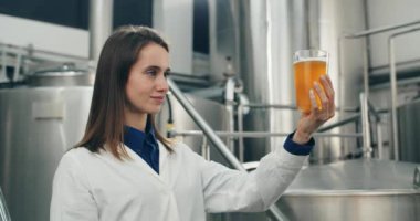 Dikkatle bakan kadın işçilerin cam bardakta taze yapılmış içeceğe bakması. Beyaz ceketli çekici bir kadın modern bira fabrikasında dikilirken bira kalitesini kontrol ediyor..
