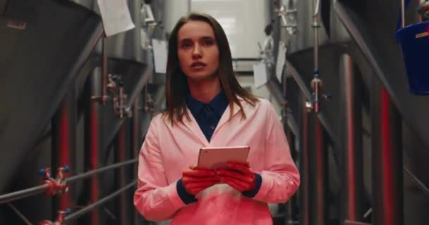 Scared female worker in white lab coat holding tablet and walking through rows of steel brewing vats. Alert signals and red lights at brewery factory. Concept of emergency situation. — Stock Video