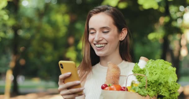 Primo piano di ragazza ridendo lodevolmente mentre guarda video su smartphone e in piedi in strada. Donna millenaria in possesso di borsa con cibo sorridente, scorrimento notizie feed. Concetto di emozioni . — Video Stock