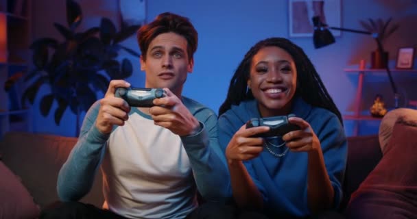 Happy couple holding wireless joysticks and play game while man loosing.Millennial people enjoing game and sitting on sofa in front of tv while woman winning. Home háttér. — Stock videók