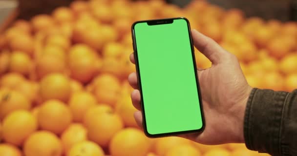 Close up view of hand holding modern mobile with template green screen in supermarket. Апельсины на размытом фоне. Концепция покупок, хрома ключ и макет . — стоковое видео