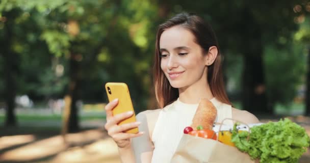 Közelkép a vonzó fiatal lány görgetés hírcsatorna, miközben tartja papírzacskó étellel. Portré gyönyörű nő segítségével smartphone és mosolygós kamera, miközben áll a parkban. — Stock videók