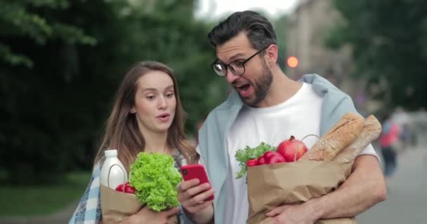 買い物や電話の画面を見て興奮した男と女が家に戻ってくる。紙袋の中に食べ物を運んで、通りを歩いている間に驚きの顔をする喜びのカップル. — ストック動画