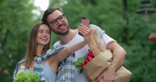 Hombre guapo y su encantadora esposa haciendo fotos o mensaje de vídeo en la cámara frontal del teléfono inteligente. Pareja alegre posando para selfies y actuando tonta con bolsas de papel con comida. Al aire libre . — Vídeos de Stock