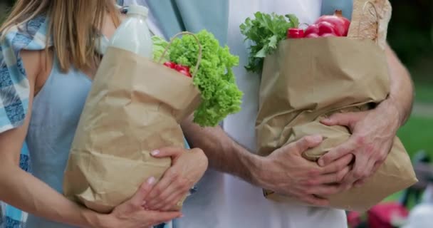 Вид сбоку на пару, несущую покупки в руках. Мужчина и женщина ходят и держат в руках бумажные пакеты, полные здоровой пищи. Концепция покупок и здорового образа жизни. На открытом воздухе . — стоковое видео