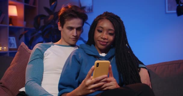 Přední pohled na mladé šťastný multietnický pár při pohledu na obrazovce smartphone, zatímco sedí na pohovce. Millennial přítel a přítelkyně procházení internetu, zatímco tráví čas spolu doma. — Stock video