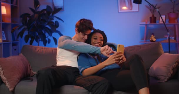 Millennial casal feliz fazendo selfies engraçadas enquanto sentado no sofá. Bonito cara e sua atraente namorada africana com dreadlocks posando ao fazer fotos e passar tempo em casa . — Vídeo de Stock