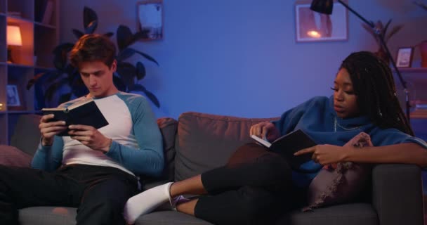 聪明的年轻夫妇在家里看书的时候会花时间。年轻的男朋友和女朋友晚上一起坐在沙发上，一边放松一边看书。休闲的概念. — 图库视频影像