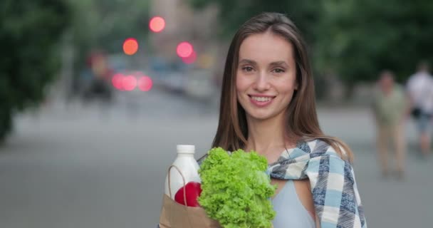 Vista da vicino della bella ragazza che gira la testa e guarda alla fotocamera mentre tiene la borsa con il cibo. Donna sorridente in posa per la fotocamera mentre in piedi in strada. Nutrizioni e vita sane . — Video Stock