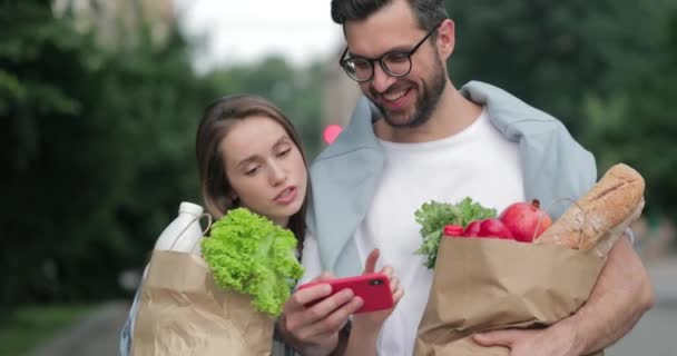 Glückliches Paar, das nach dem Einkaufen nach Hause kommt und Lebensmittel in Papiertüten trägt. Fröhliche Frau und Mann suchen und berühren Smartphone-Bildschirm im horizontalen Modus und zu Fuß auf der Straße. — Stockvideo