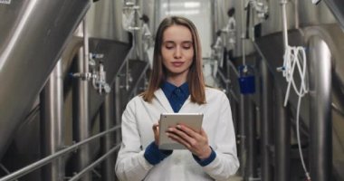 Beyaz laboratuvar önlüklü bir kadının portresi bira fabrikasındaki tablete veri giriyor ve kameraya bakıyor. Kadın işçiler içecek ekipmanlarının yanında dururken dijital ekrana dokunup yazıyorlar..