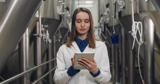描述穿着白色实验室外套的女性在酿酒厂的平板电脑上输入数据，并看着相机。女工站在饮料设备旁边触摸和打字数字显示. — 图库视频影像