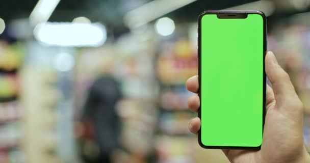 Vista de perto da mão segurando smartphone moderno com tela simulada no modo vertical. Conceito de chave chroma e tela verde. Fundo de supermercado desfocado. Interior . — Vídeo de Stock