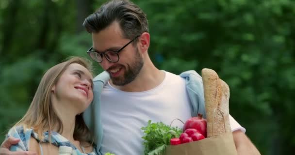 Närbild av härliga par kommunicera och titta på varandra när du går. Glad man i glasögon omfamnar sin vackra fru medan de bär papperspåse med mat. Utomhus. — Stockvideo