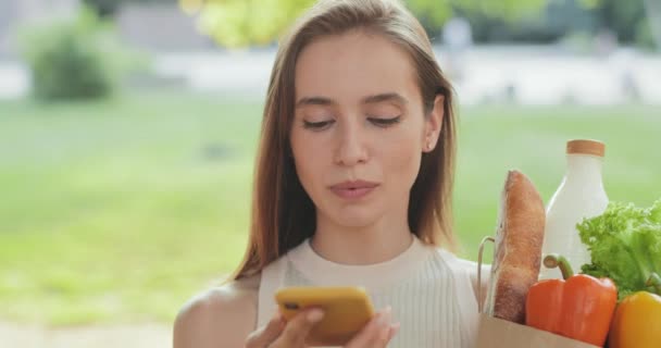 Close up de mulher milenar falando ao usar telefone assistência ai móvel e sorrindo. Menina bonita ditando mensagem de voz enquanto segurando saco de papel com mantimentos ao ar livre . — Vídeo de Stock