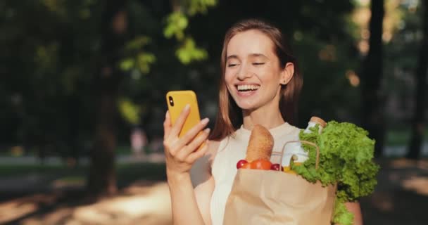 Χαρούμενη κοπέλα που γελάει εγκωμιαστικά, ενώ διαβάζει αστεία μηνύματα στο smartphone της και στέκεται στο δρόμο. Millennial όμορφη γυναίκα κρατώντας τσάντα με παντοπωλεία χαμογελώντας ενώ κύλιση ειδήσεων ζωοτροφών. — Αρχείο Βίντεο