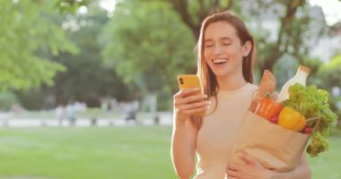 Akıllı telefon ekranına bakıp gülen neşeli genç bir kadın. Güzel bir milenyum kızı, elinde kese kağıdıyla durup telefonunu kullanırken gülümsüyor. Yaşam tarzı kavramı.