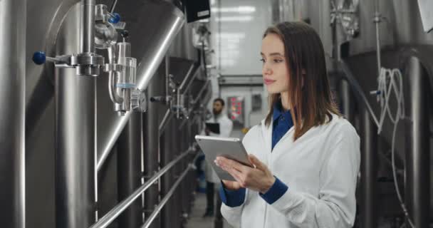 Vista de la cosecha de buen aspecto trabajador femenino que toca la pantalla al introducir datos en la tableta. Mujer y hombre con bata de laboratorio blanca comprobando los medidores del equipo y de pie en filas de cubas de cerveza . — Vídeos de Stock