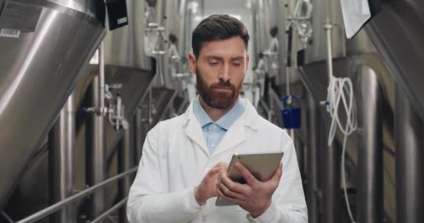 Retrato del hombre con bata blanca ingresando datos en la tableta en la cervecería y mirando a la cámara. Trabajador masculino tocando y escribiendo la pantalla digital mientras que está parado cerca del equipo de la bebida . — Vídeos de Stock