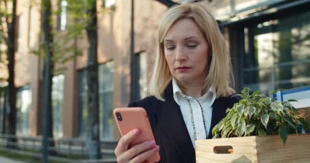 Primer plano de empleada en ropa formal sosteniendo caja con sus pertenencias mientras está de pie en la calle. Mujer desplazándose por la pantalla del teléfono móvil mientras busca un nuevo trabajo después de ser despedido . — Vídeos de Stock