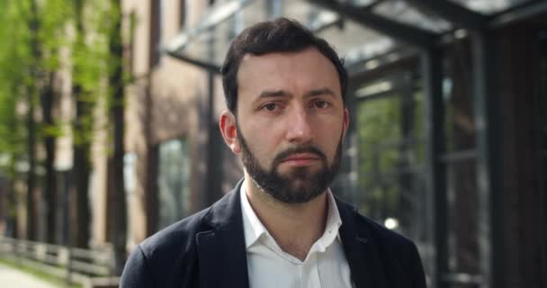 Vista de perto do homem desapontado nos anos 30 virando a cabeça e olhando para a câmera. Retrato de homem barbudo trabalhador de escritório de terno em pé na rua perto do edifício moderno. Conceito de negócio . — Vídeo de Stock