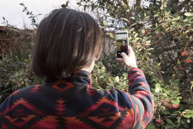 Genç adam cep telefonuyla kırmızı böğürtlenlerin yeşil çalılarının önünde fotoğraf çekiyor.