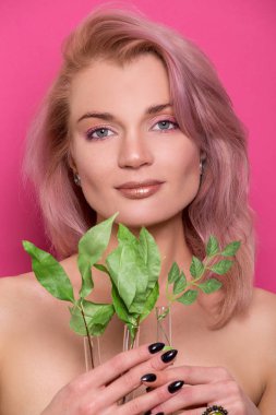 Elinde yeşil yapraklı bir dal tutan güzel Avrupalı model bir kızın üzerinde parlak altın bir makyaj. Fotoğraf stüdyosunun pembe arka planında izole edilmiş.