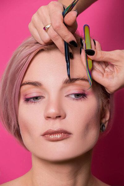 Крупный план красивой девушки европейского вида держит в руке пинцет для бровей рядом с лицом в студии изолирован на розовом фоне
