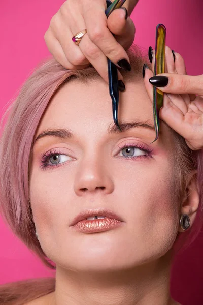 Close-up of a beautiful girl of European appearance holds in her hand tweezers for eyebrows near the face in the studio isolated on a pink background