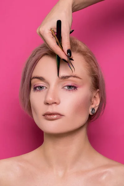 Close-up of a beautiful girl of European appearance holds in her hand tweezers for eyebrows near the face in the studio isolated on a pink background
