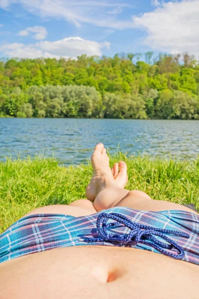 Man with checkered bathing slip and bare feet at lake / river, meadow / grass — Stock Photo, Image