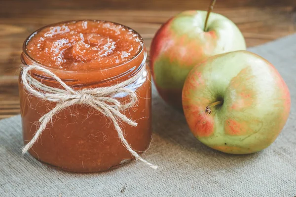 Apple jam in bank with apples, slice of bread with jam and spoon of jam on linen fabric on wooden background.