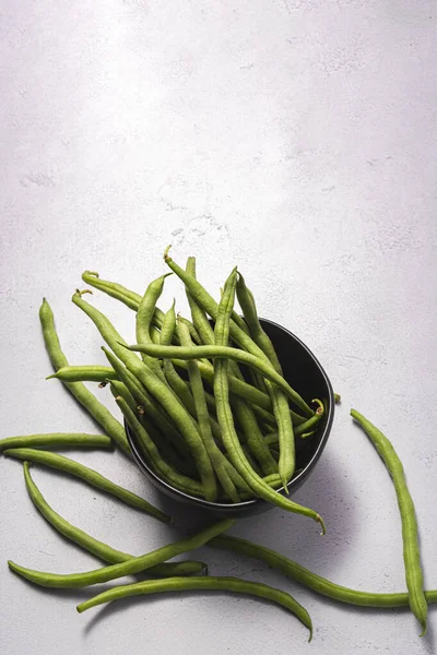 Fresh raw green beans in a black bowl on an textured white surface.