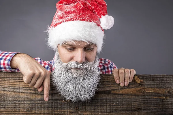 一个英俊的男人的特写镜头 长胡子和胡子与红色的圣诞老人帽子与毛皮显示的东西下来的灰色 免版税图库图片