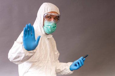 Cerrahi maskeli, gözlüklü ve koruyucu elbiseli bir doktor telefonunu tutuyor ve gri arka planda durduğunu gösteriyor..