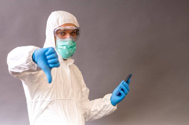 Cerrahi maskeli, gözlüklü ve koruyucu elbiseli bir erkek doktor. Telefonunu tutuyor ve gri arka plana işaret ediyor..