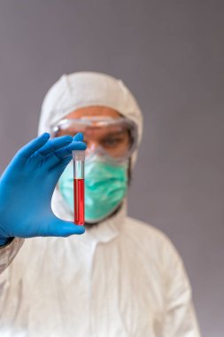 Cerrahi maskeli, gözlüklü ve koruyucu elbiseli bir erkek doktor gri arka planda kan örnekleri tutuyor. İlaç şişeleri Covid-19.