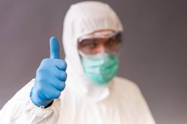 Cerrahi maskeli, gözlüklü ve koruyucu elbiseli bir erkek doktor gri arka planı işaret ediyor..