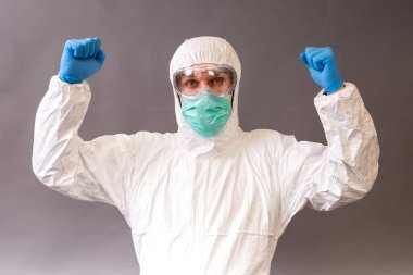 Cerrahi maskeli, gözlüklü ve gri arka planda koruyucu elbiseli bir erkek doktor..