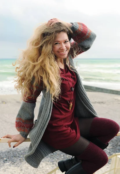 Vackra glada leende kvinna porträtt sitter på ett räcke mot hösten havet, långa hårstrån, kort mode mörk röd klänning och grått jersey — Stockfoto