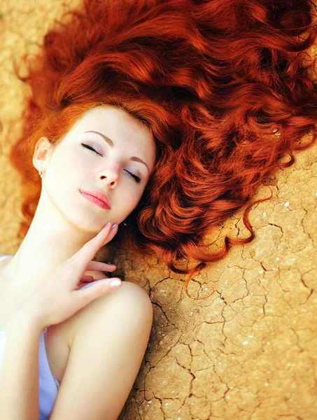 Beautiful young woman portrait with long curly rude hair and perfect fresh skin lying on the dried up ground with cracks — Stock Photo, Image