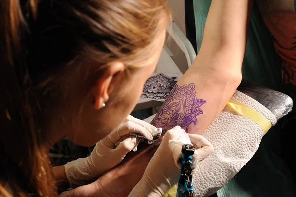 Young cute woman master tattooist makes the tattoo on a hand under the lamp light — Stock Photo, Image