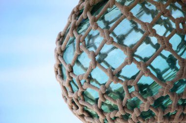 Turquoise green sea glass sphere tied up with nautical rope & knots against a blue sky.  clipart